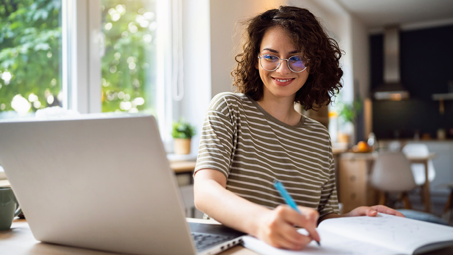 Woman smiling and writing notes in front of a laptop at home.