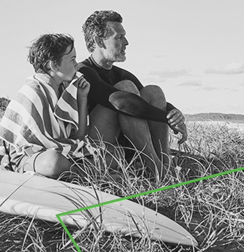 Boy and man sitting on the beach next to a surfboard, looking out at the ocean.