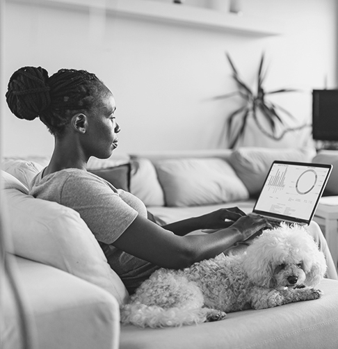 Woman sitting on a couch next to her dog, looking at a laptop screen showing investment charts. 